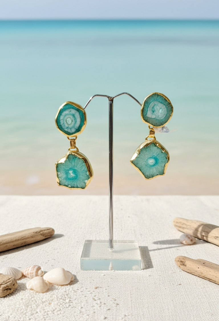 Pair of gold-plated, double dangle beach wear earrings featuring raw, polished aqua blue/teal druzy agate stones displayed on a stand with a tropical beach and ocean background