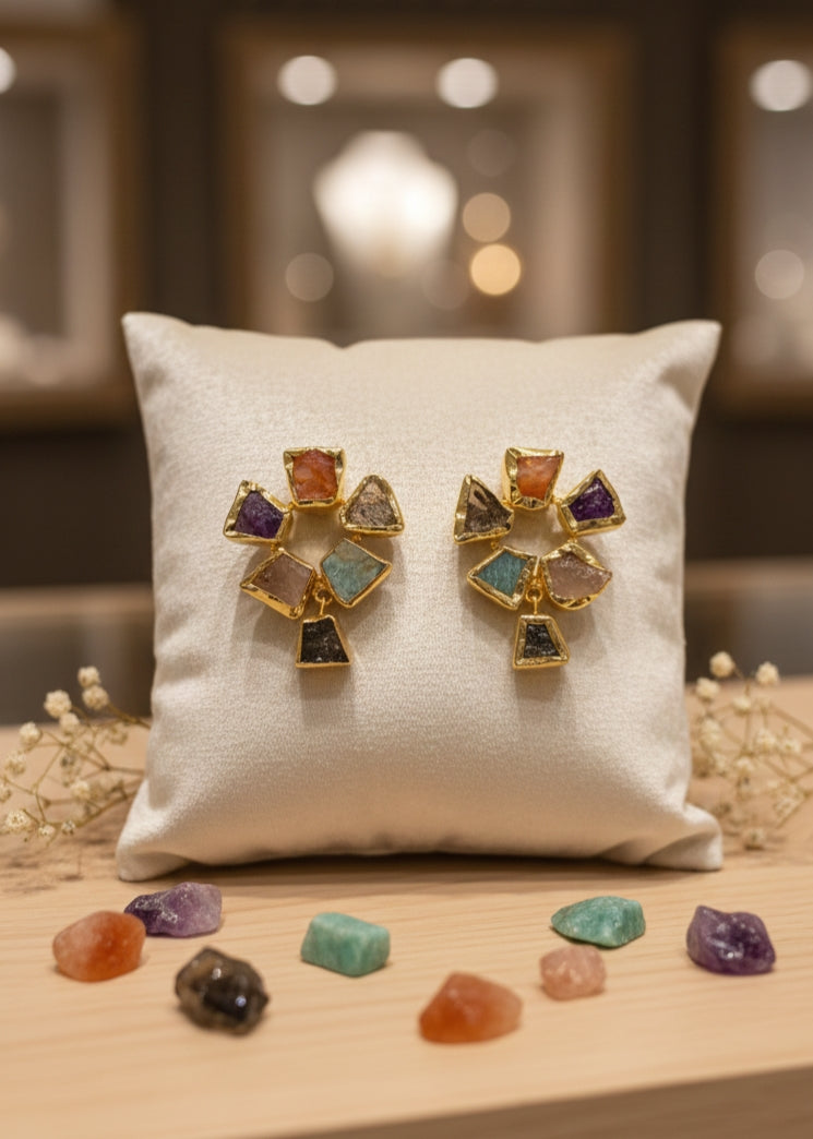 Gold statement earrings featuring multi-colored raw gemstones arranged in a fan shape, displayed on a white jewelry cushion.